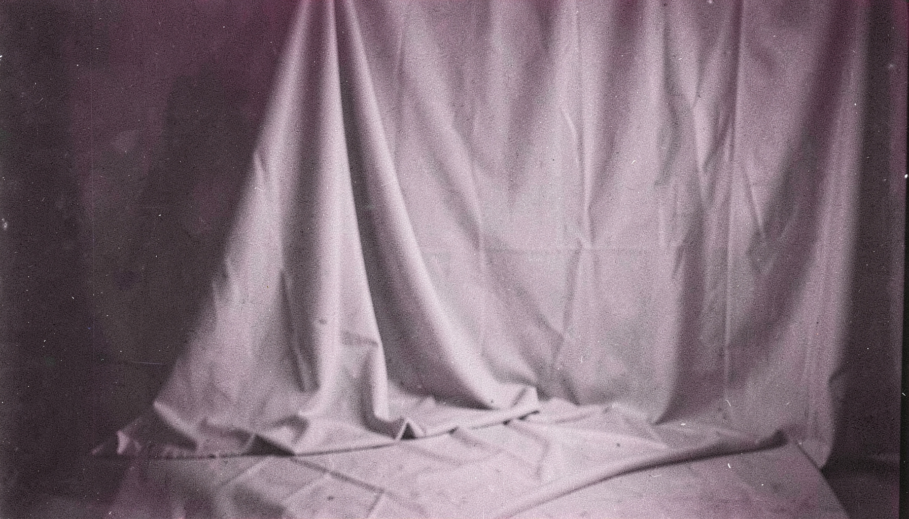 Black and white film photograph with visible grain showing a softly lit studio interior with draped fabric and subtle shadows, contemplative and atmospheric mood
