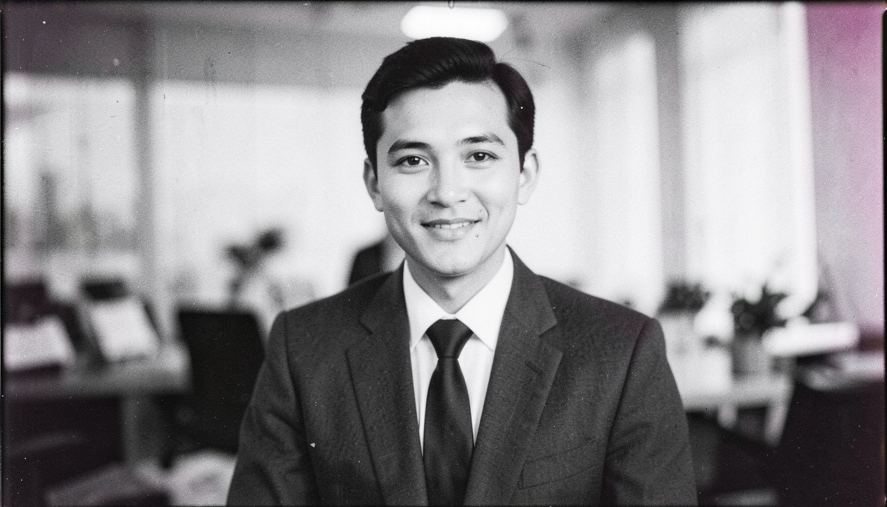 Analog black-and-white photograph with visible film grain, a business portrait of a person in tailored clothing against a softly blurred office backdrop, confident and polished mood