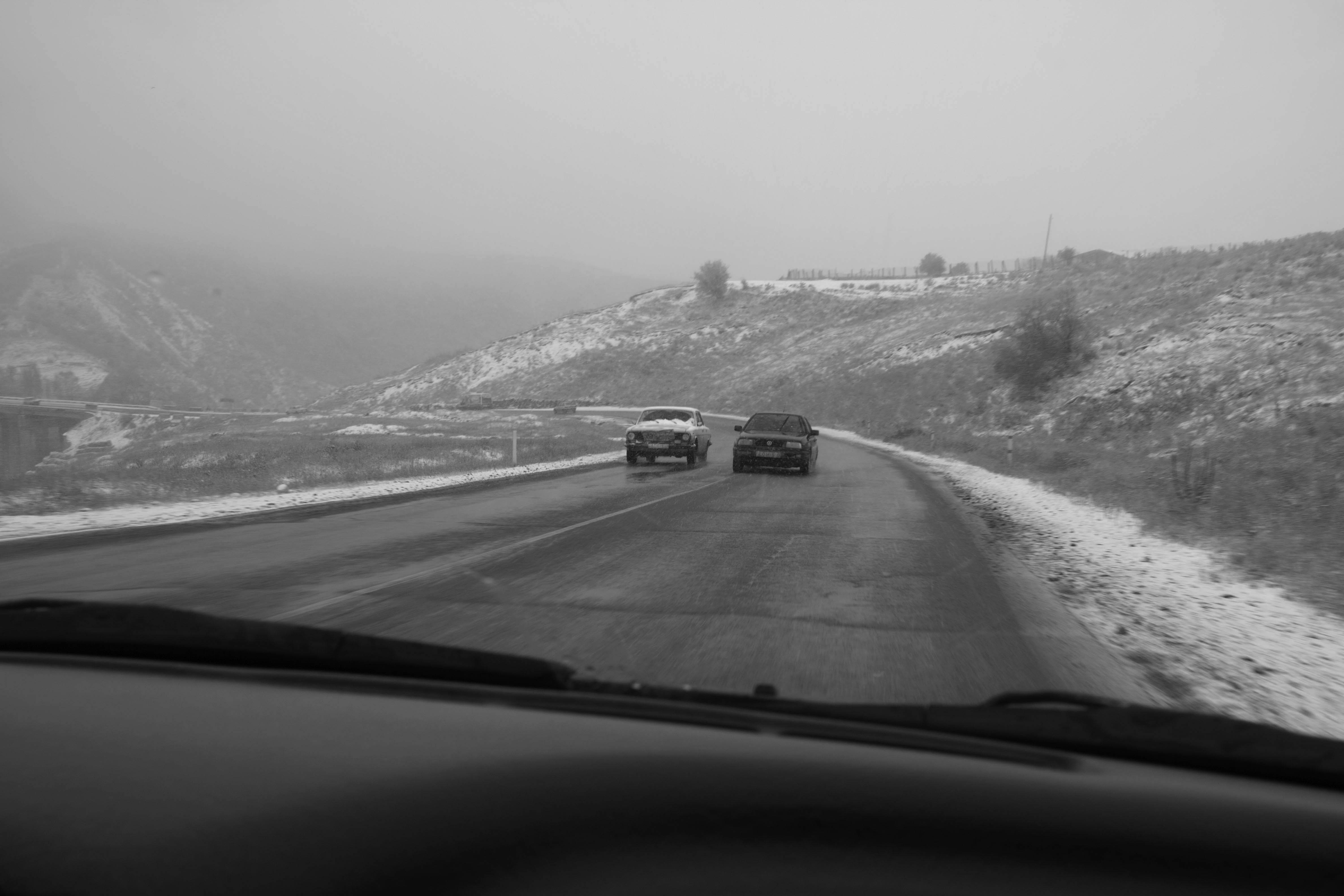 Black-and-white view through a car windshield of two vehicles driving on a snowy mountain road, overcast mood